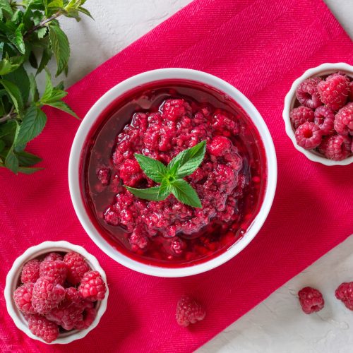 Delicious raspberry jam in a bowl, fresh berries, mint leaves on a pink napkin. Top view.