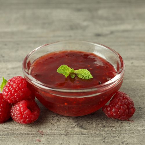 Glass bowl of raspberry jam with ingredients on gray textured background